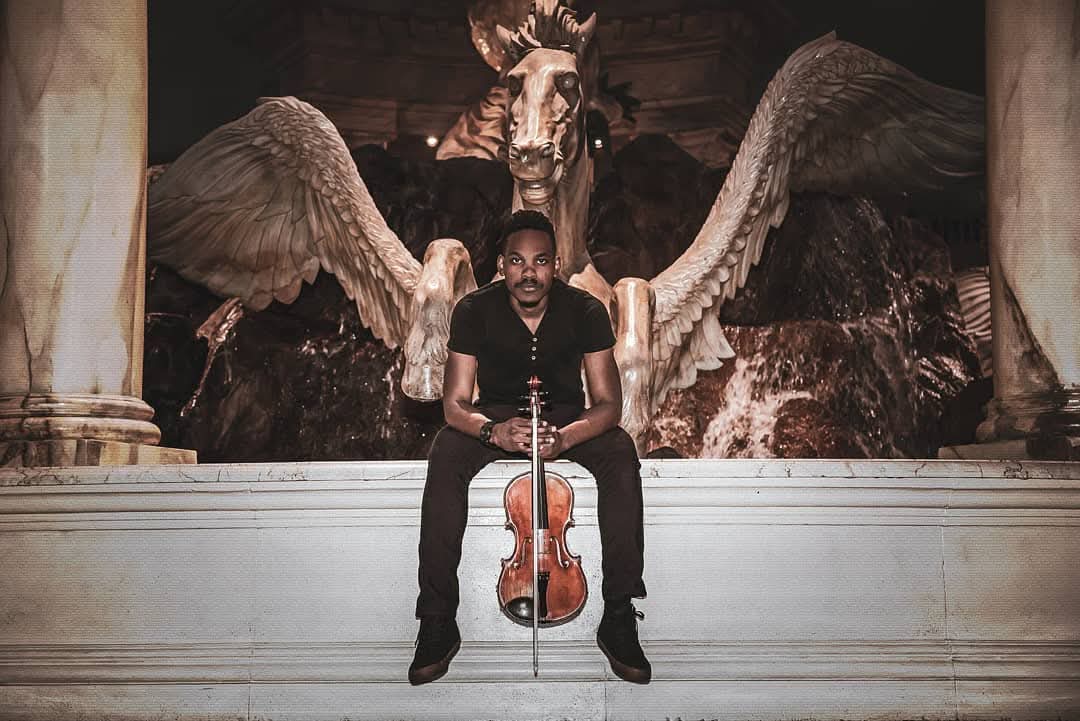 Rahmaan Phillip seated at the winged-horse fountain in Las Vegas, violin upright, looking to camera.